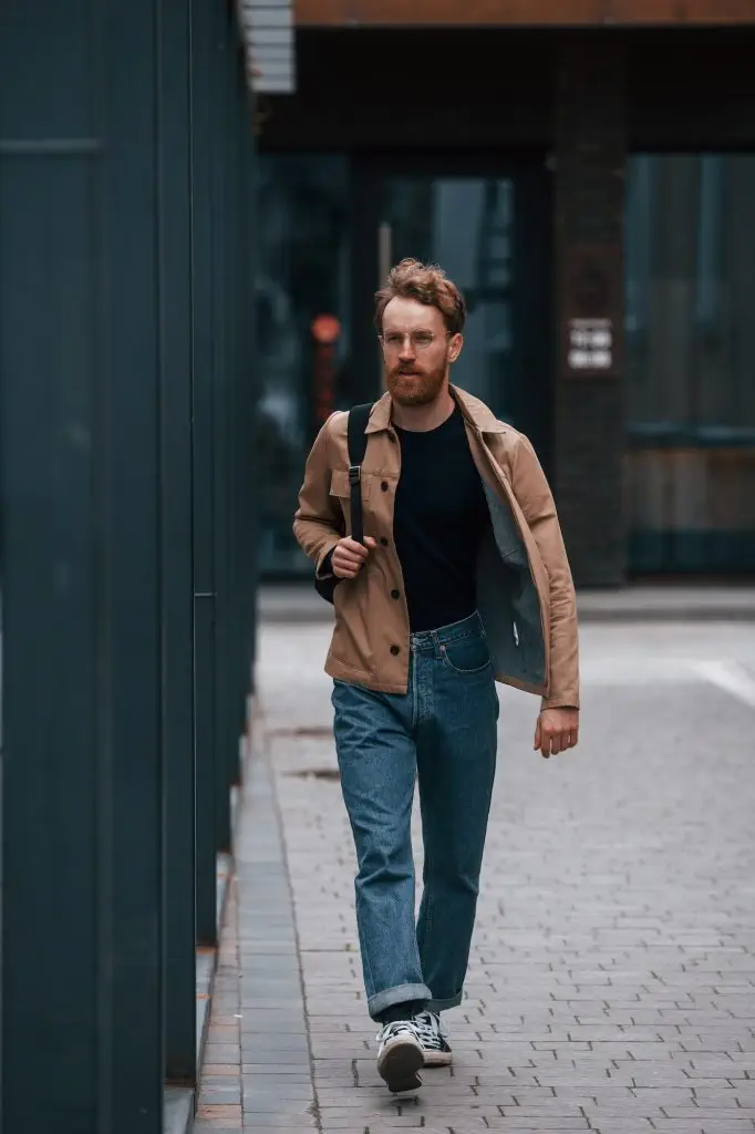 Front view. Stylish man with beard in khaki colored jacket and in jeans is outdoors near building