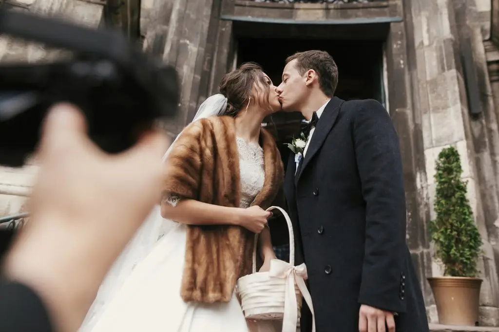 photographer taking photo of gorgeous bride in coat and stylish groom near church