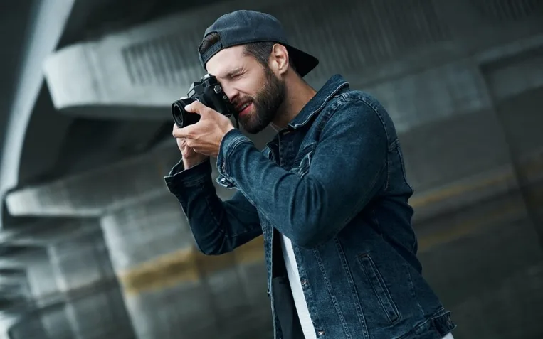 Photography hobby. Young stylish man standing on city street taking photos on camera concentrated